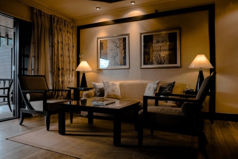 A cozy, dimly lit hotel suite living area with dark wood floors and a light area rug. A beige sofa with decorative pillows is flanked by two dark wood armchairs and matching lamps with white shades. Two framed abstract floral artworks hang above the sofa on a beige wall with dark accent trim. A glass door opens to a balcony with sheer and patterned drapes drawn across.