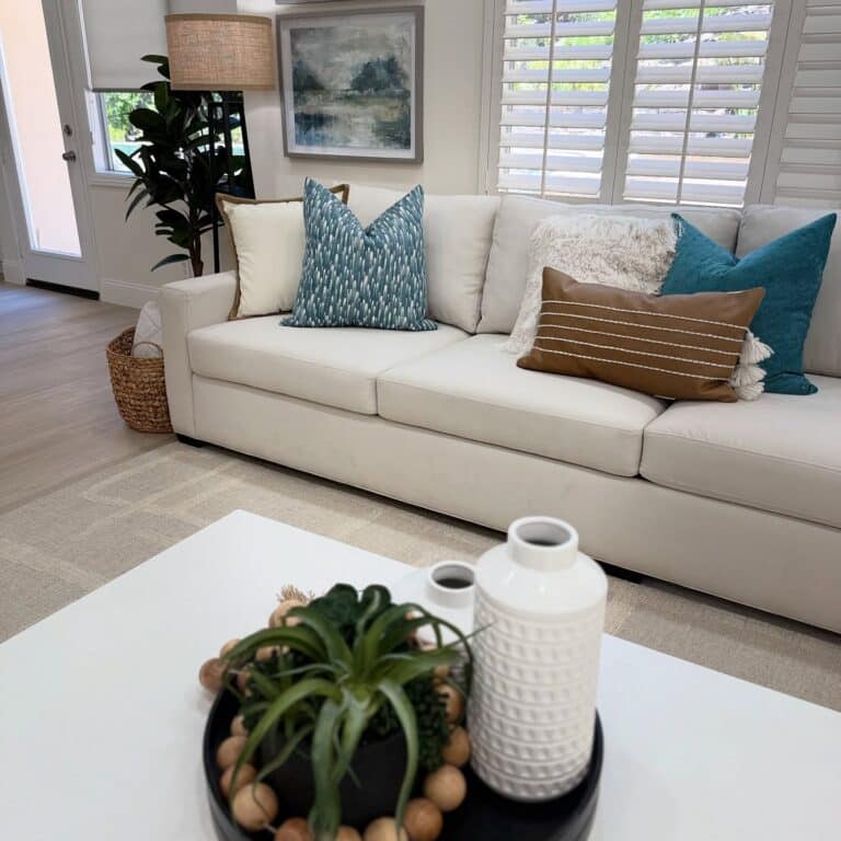 A cozy living room with an off-white sectional sofa adorned with blue, brown, and cream pillows. Below a window with white plantation shutters, a large botanical print hangs on the wall above the couch.