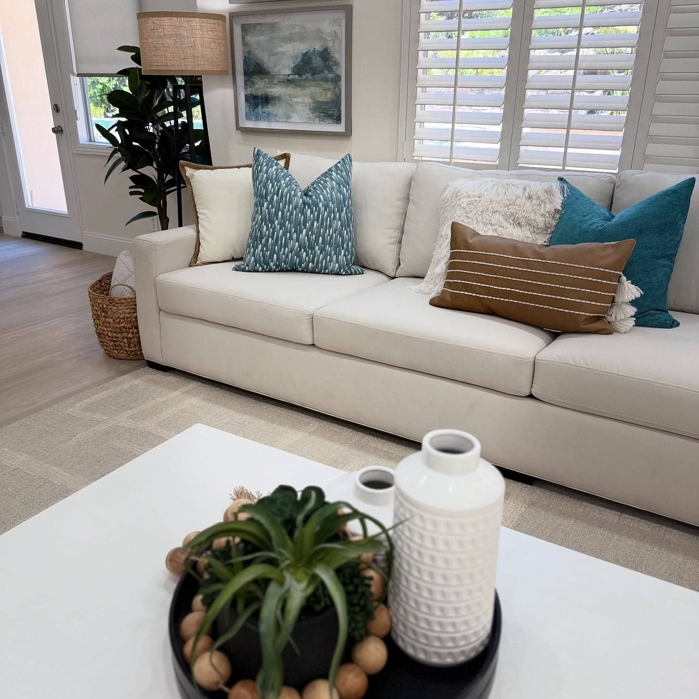 Blog 4 A cozy living room with an off-white sectional sofa adorned with blue, brown, and cream pillows. Below a window with white plantation shutters, a large botanical print hangs on the wall above the couch.